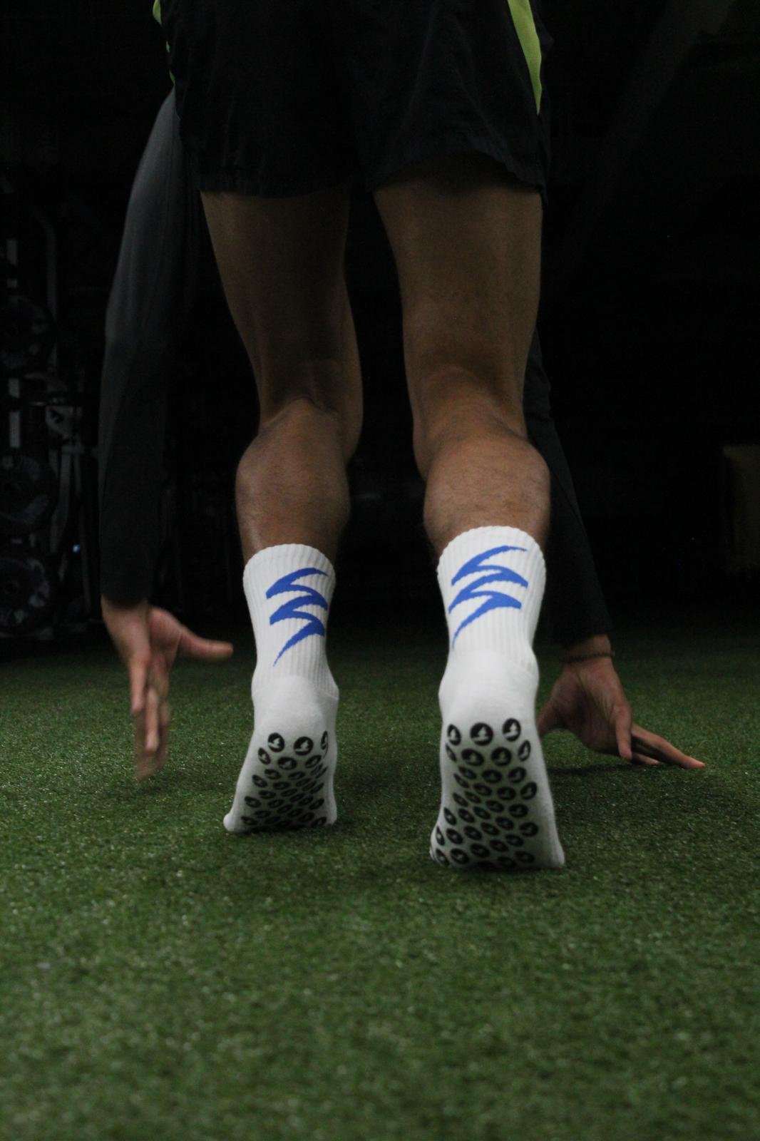 An athlete viewed from behind in a sprint-start stance on training turf, wearing white ZIP performance socks with the bolt mark in royal blue and the grip-dot sole visible